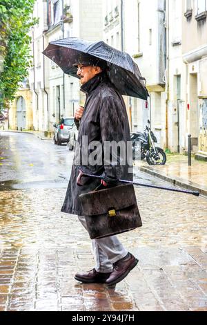 Älterer Geschäftsmann mit Aktentasche, Gehstock und Regenschirm, der bei strömendem Regen auf der Stadtstraße spaziert - Tours, Indre-et-Loire (37), Frankreich. Stockfoto