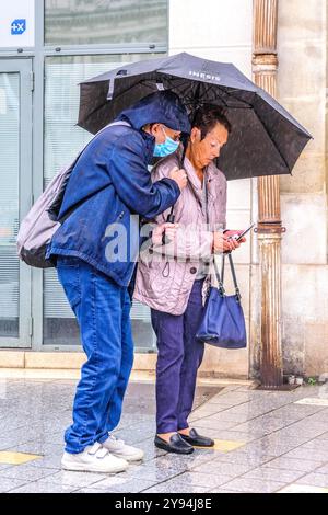 Älteres orientalisches Paar unter Regenschirm im strömenden Regen mit Blick auf das Mobiltelefon - Tours, Indre-et-Loire (37), Frankreich. Stockfoto
