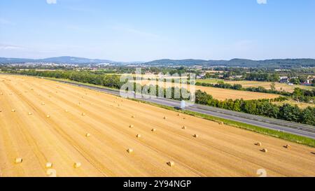 Aus der Vogelperspektive zeigt ein Feld mit Heuballen neben einer Autobahn, umgeben von grünen Bäumen und fernen Hügeln unter einem klaren blauen Himmel. Stockfoto