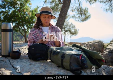 Eine Frau sitzt auf einer felsigen Oberfläche umgeben von Natur und genießt eine ruhige Pause im Freien. Sie überprüft ihr Handy, mit Reiseutensilien und einer Thermoskanne Stockfoto