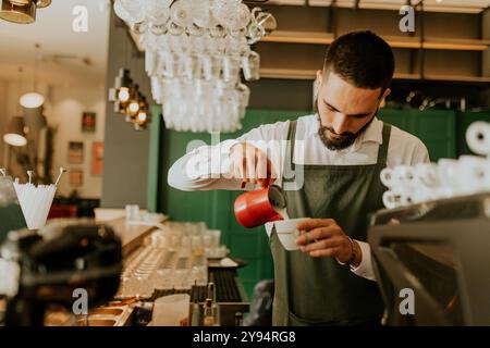 In einem warmen und einladenden Café gießt ein engagierter Barista fachmännisch Milch in eine Kaffeetasse und sorgt so für ein angenehmes Erlebnis für Gäste. Stockfoto