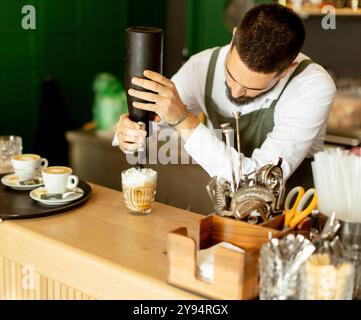 Ein engagierter Barista dekoriert einen Dessertkaffee elegant mit Schlagsahne und zeigt seine Kunst in einem lebhaften Café mit warmem Licht und Invit Stockfoto