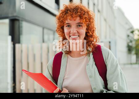 Ein lächelnder rothaariger Student, der in die Kamera blickt, während er eine rote Mappe hält und einen Rucksack trägt. Er steht draußen in einer urbanen Umgebung, zeigt Selbstvertrauen und eine positive Einstellung an einem hellen Tag. Hochwertige Fotos Stockfoto