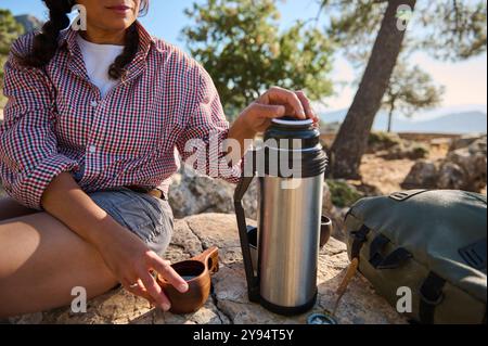 Eine Frau genießt ein friedliches Picknick im Freien an einem sonnigen Tag, sitzt auf felsigem Gelände mit einer Thermoskanne und einer Tasse. Bäume und Berge sind im Hintergrund zu sehen Stockfoto