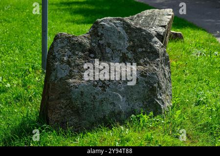 Ein großer, moosbedeckter Felsen befindet sich inmitten von üppigem grünem Gras neben einem Weg, der sich im Sonnenlicht in einer friedlichen Parkumgebung sonnt und ein natürliches aest bietet Stockfoto