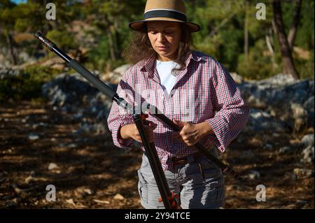 Frau in lässiger Outdoor-Kleidung, die während des sonnigen Tages ein Stativ vor einem Naturparkhintergrund aufstellt. Stockfoto