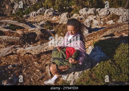 Eine Frau genießt einen friedlichen Moment in der Natur, sitzt auf einem Felsen, während sie ihren Rucksack während einer Wanderung in einer zerklüfteten, felsigen Landschaft, umgeben von Tre, überprüft Stockfoto