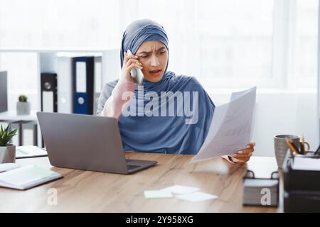 Betonte muslimische Geschäftsfrau, die die Dokumentation überprüft und im Büro per Handy spricht Stockfoto