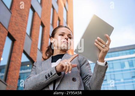 Netzwerke und Unternehmensunternehmen: Aus einem niedrigen Winkel sehen Sie eine selbstbewusste Geschäftsfrau, die außerhalb eines Bürogebäudes an einem digitalen Tablet arbeitet. Stockfoto