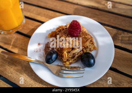 Ein köstlich aussehendes Stück Orangenkuchen sitzt auf einem weißen Teller, mit einer Himbeere geschmückt und von Heidelbeeren umgeben. Ein Glas Orangensaft Stockfoto