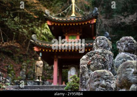 Eine Gruppe von moosbedeckten Steinstatuen steht vor einem traditionellen Tempel in einer Waldlandschaft und schafft eine ruhige und historische Atmosphäre. Stockfoto