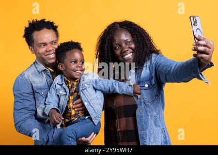 Stolze glückliche Eltern machen gerne Fotos mit ihrem niedlichen Kleinkind vor gelbem Hintergrund und fangen lustige Momente zusammen ein. Fröhliches afroamerikanisches Paar posiert für Selfies mit dem Sohn. Stockfoto