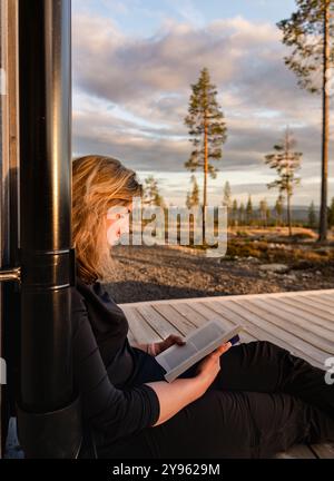Eine schöne Frau sitzt friedlich auf einem Deck und liest während der goldenen Stunde ein Buch mit Bergen und Bäumen im Hintergrund, das die Natur umschmeichelt Stockfoto