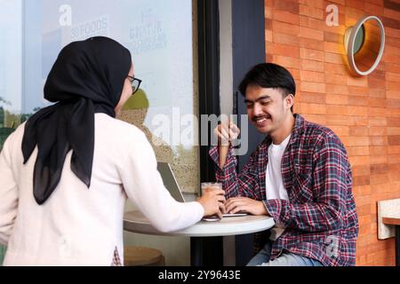 Der junge Indonesisch-asiatische Lächeln seiner Freundin zu, die Getränke serviert, während er bei Cafe Table, WFC Concept arbeitet Stockfoto