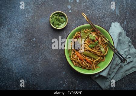 Asiatisch gebratene udon-Nudeln mit Huhn, Gemüse, Pilzen in grüner Keramikschüssel mit Holzstäbchen auf rustikalem dunkelblauem Betonhintergrund f Stockfoto