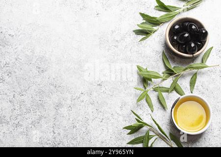 Schwarze Oliven und extra natives Olivenöl in kleiner Schüssel mit Olivenbaumzweigen von oben Blick auf weißen Betonsteinhintergrund, Kopierraum, Lebensmittel-Fotogra Stockfoto