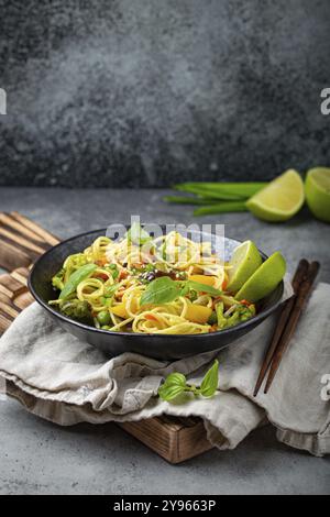 Asiatische vegetarische Nudeln mit Gemüse und Limette in schwarzer rustikaler Keramikschale, Holzstäbchen auf Schneidebrett, Winkelansicht auf Steinhintergrund. Coo Stockfoto