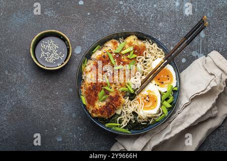 Asiatische Nudeln Ramen-Suppe mit frittiertem Panko-Hähnchenfilet und gekochten Eiern in einer Keramikschüssel mit Koteletts und Sojasauce auf rustikalem Backgroun aus Stein Stockfoto