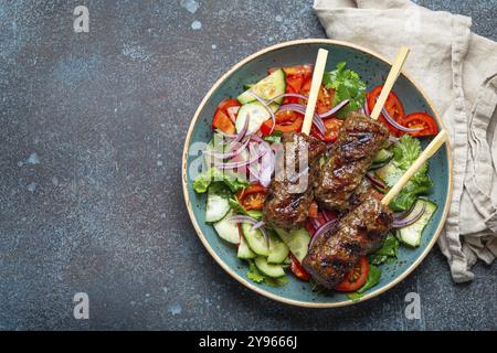 Gegrilltes Spießfleisch Rindfleisch Kebabs auf Stäbchen serviert mit frischem Gemüsesalat auf Teller auf rustikalem Betonhintergrund von oben. Traditionelles Mittleres Eas Stockfoto