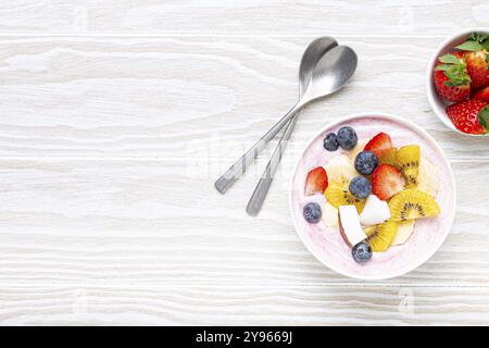 Gesundes Frühstück oder Dessert Joghurt Schüssel mit frischer Banane, Erdbeere, Heidelbeere, kokos, Kiwi Top View auf rustikalem hölzernen weißen Hintergrund mit Löffel. Stockfoto