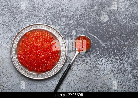 Kleiner Metallteller mit rotem Lachskaviar und einem Löffel von oben auf grauem Betonhintergrund, festliche Luxus-Delikatesse und Vorspeise, Lebensmittelfotografie, fo Stockfoto