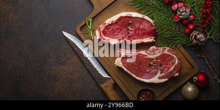 Zwei rohe Rindersteaks mit rohem Fleisch auf Holzbrett mit Messer und Gewürzen auf dunklem rustikalem Hintergrund mit festlicher Weihnachtsdekoration von A Stockfoto