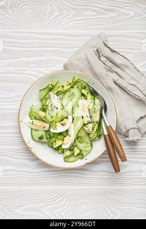 Gesunde grüne Avocadosalatschale mit gekochten Eiern, Gurken, Edamame Bohnen, Olivenöl und Kräutern auf Keramikplatte, Blick von oben auf weißen Holzrost Stockfoto