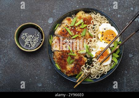 Asiatische Nudeln Ramen-Suppe mit frittiertem Panko-Hähnchenfilet und gekochten Eiern in einer Keramikschüssel mit Koteletts und Sojasauce auf rustikalem Backgroun aus Stein Stockfoto