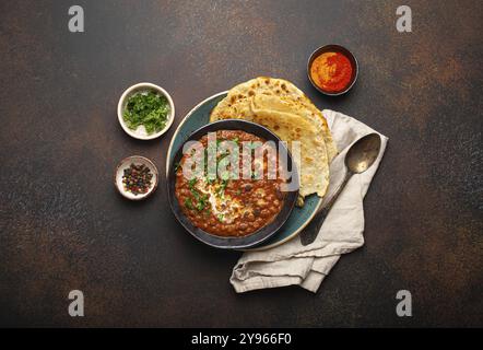 Traditionelles indisches Punjabi-Gericht Dal Makhani mit Linsen und Bohnen in schwarzer Schüssel serviert mit Naan-Fladenbrot, frischem Koriander und zwei Löffeln auf braunem C Stockfoto