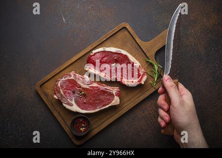 Männliche Hand hält Messer über zwei rohe, rohe, rohe, rohe, ungekochte, marmorierte Steaks mit Rinderrippen auf Holzbrettern und Würzmittel auf dunklem rustikalem Hintergrund Stockfoto