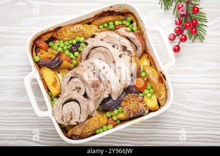 Festliches weihnachtliches, gerolltes Schweineschneidfleisch in weißen Auflaufform mit Kartoffeln, Gemüse und Kräutern auf rustikalem weißem hölzernem Hintergrund von oben gebraten. B Stockfoto