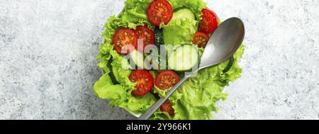 Gesunder Gemüsesalat mit frischen Gurken, Tomaten, frischen Eisbergsalatblättern in einer Schüssel auf rustikalem weißem Beton mit einem Löffel. Ve Stockfoto