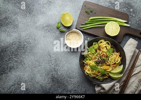 Asiatische vegetarische Nudeln mit Gemüse und Limette in schwarzer rustikaler Keramikschüssel, Holzstäbchen, Schneidebrett mit gehackter grüner Zwiebel von oben Stockfoto