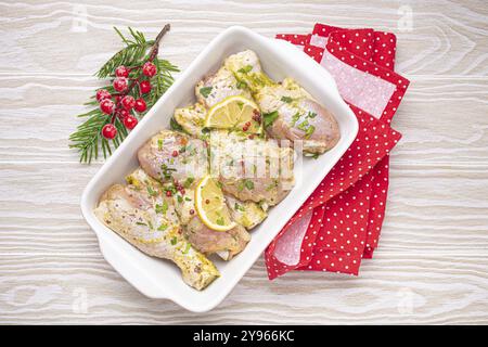 Zubereitung festlicher Mahlzeit am Heiligabend mit marinierten Hähnchenstangen. Rohe Hähnchenschenkel mit Kräutern, Zitrone in weißem Keramikauflauf, Blick von oben auf Ligh Stockfoto