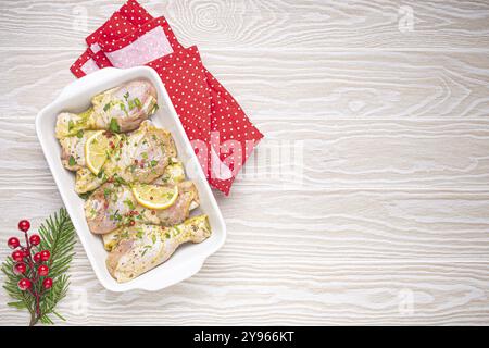 Zubereitung festlicher Mahlzeit am Heiligabend mit marinierten Hähnchenstangen. Rohe Hähnchenschenkel mit Kräutern, Zitrone in weißem Auflauf, Blick von oben auf Holzküche Stockfoto