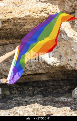 Eine Regenbogenfahne wird vor einem felsigen Hintergrund unter einem sonnigen Himmel gehalten, Frau mittleren Alters, im Freien winkende Stolzfahne, Konzept von Libertat, Equali Stockfoto