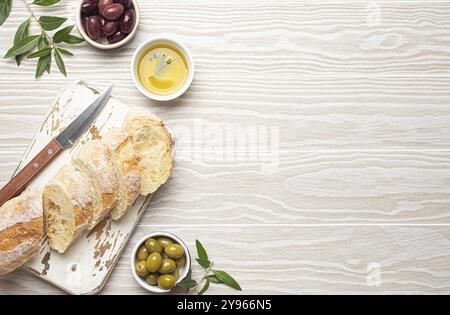 In Scheiben geschnittene frische Ciabatta auf Schneidebrett, grüne und braune Oliven, Olivenöl mit Rosmarin, Olivenzweige, weißer hölzerner rustikaler Hintergrund t Stockfoto