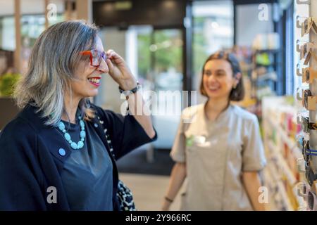 Eine reife Frau, die im Optikgeschäft neben der Verkäuferin eine Brille anprobiert Stockfoto