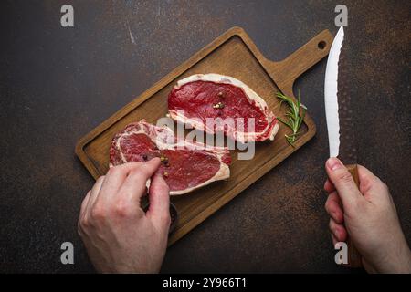 Männliche Hand hält Messer über zwei rohe, rohe, rohe, rohe, ungekochte, marmorierte Steaks mit Rinderrippen auf Holzbrettern und Würzmittel auf dunklem rustikalem Hintergrund Stockfoto