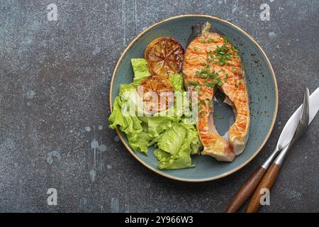 Gegrilltes Fisch Lachssteak und grüner Salat mit Zitrone auf Keramikplatte auf rustikalem blauem Stein Hintergrund Draufsicht, ausgewogene Ernährung oder gesunde Ernährung Mea Stockfoto