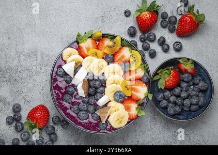 Gesunder Sommer Acai Smoothie Bowl mit Chiasamen, frischer Banane, Erdbeere, Heidelbeere, kokos, kiwi-Blick von oben auf rustikalem Betonhintergrund mit Löffel Stockfoto