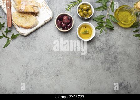 Komposition mit geschnittenem frischen Ciabatta auf Schneidebrett, grünen und braunen Oliven, Olivenöl mit Rosmarin, Olivenzweigen auf grauem Betonstein r Stockfoto