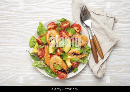 Gesunder Salat mit gegrillten Garnelen, Avocado, Kirschtomaten und grünen Blättern auf weißem Teller mit Besteck auf weißem hölzernem rustikalem Hintergrund von oben. Stockfoto