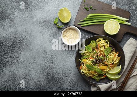 Asiatische vegetarische Nudeln mit Gemüse und Limette in schwarzer rustikaler Keramikschüssel, Holzstäbchen, Schneidebrett mit gehackter grüner Zwiebel von oben Stockfoto