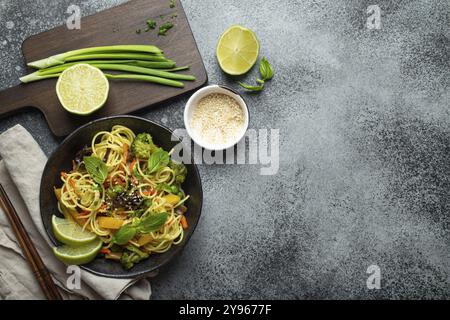 Asiatische vegetarische Nudeln mit Gemüse und Limette in schwarzer rustikaler Keramikschüssel, Holzstäbchen, Schneidebrett mit gehackter grüner Zwiebel von oben Stockfoto