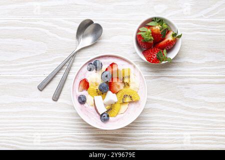 Gesundes Frühstück oder Dessert Joghurtschale mit frischer Banane, Erdbeere, Heidelbeere, kokos, Kiwi-Top-Blick auf rustikalen hölzernen weißen Hintergrund mit Löffel, Stockfoto