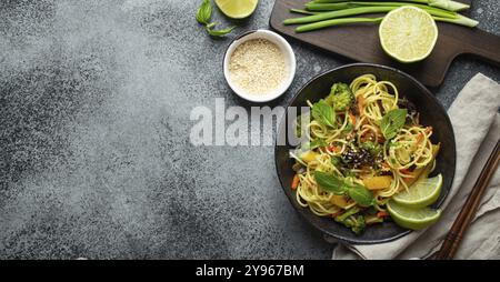 Asiatische vegetarische Nudeln mit Gemüse und Limette in schwarzer rustikaler Keramikschüssel, Holzstäbchen, Schneidebrett mit gehackter grüner Zwiebel von oben Stockfoto