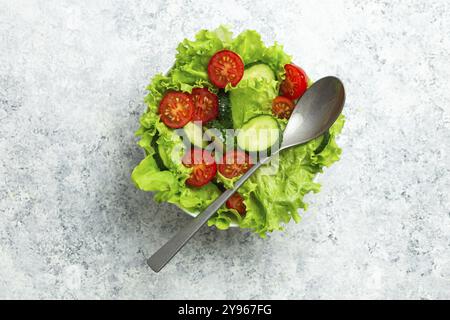 Gesunder Gemüsesalat mit frischen Gurken, Tomaten, frischen Eisbergsalatblättern in einer Schüssel auf rustikalem weißem Beton mit einem Löffel. Ve Stockfoto