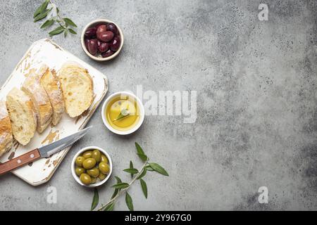 In Scheiben geschnittene frische Ciabatta auf Schneidebrett, grüne und braune Oliven, Olivenöl mit Rosmarin, Olivenzweige auf grauem Betonstein rustikale bac Stockfoto