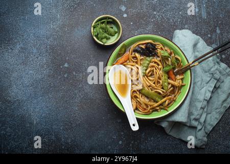 Asiatische Suppe mit udon-Nudeln, Huhn und Gemüse in grüner Keramikschüssel mit Holzstäbchen auf rustikalem dunkelblauem Betonhintergrund von oben C. Stockfoto